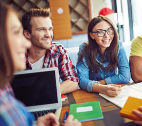 Junge Erwachsene arbeiten gemeinsam an einem Tisch, lachen und besprechen Notizen. Ein Laptop und Notizbücher liegen auf dem Tisch.“
